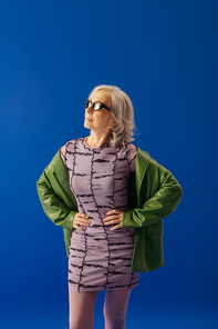 stylish senior model in sunglasses and purple dress with green leather jacket standing with hands on waist isolated on blue