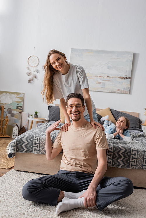 woman leaning on shoulders of happy husband sitting on carpet near daughter on bed