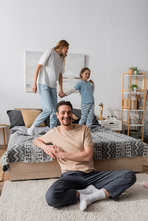 happy bearded man sitting on carpet near wife and daughter on bed on blurred background