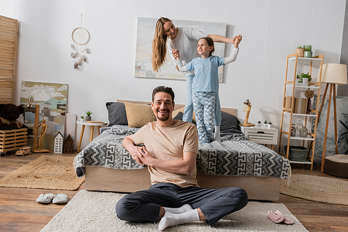 cheerful bearded man sitting on carpet near wife and daughter standing on bed on blurred background