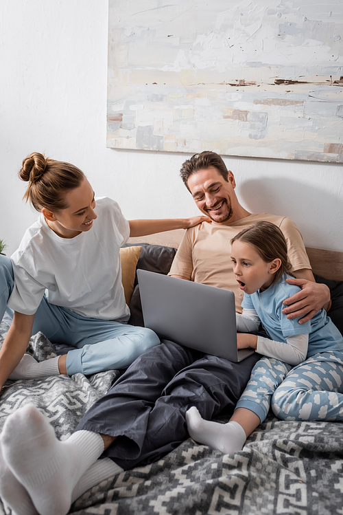 cheerful parents and shocked kid watching movie on laptop in bedroom