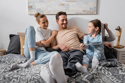 cheerful parents looking at happy kid holding laptop in bedroom