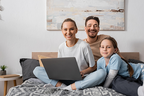 smiling parents and happy child looking at camera near laptop in bedroom