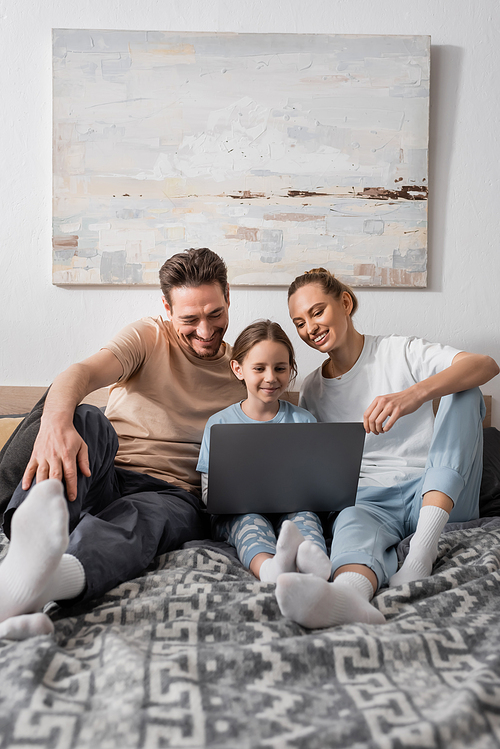 cheerful parents and happy daughter watching movie on laptop in bedroom
