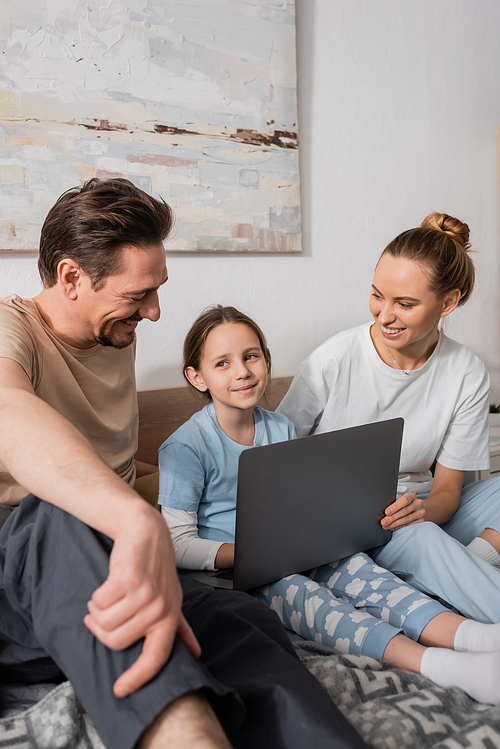 cheerful parents looking at happy kid holding laptop and smiling in bedroom
