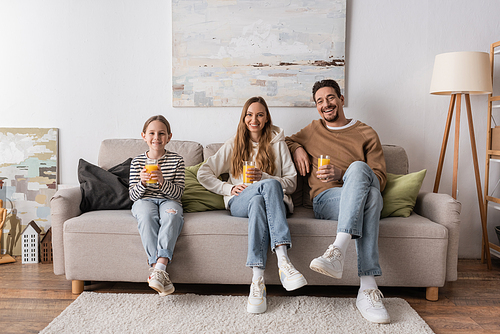full length of cheerful family holding glasses of orange juice and sitting on sofa