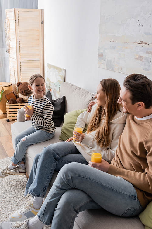 cheerful parents holding glasses of orange juice and looking at happy daughter while sitting on sofa