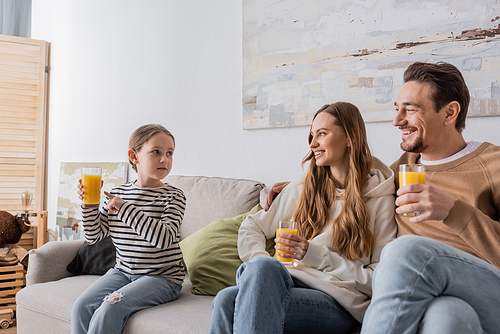 cheerful parents holding glasses of orange juice and looking at daughter while sitting on couch