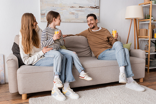 full length of cheerful daughter sitting on laps of mother and holding glass of orange juice near positive father