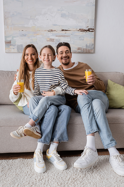 full length of cheerful daughter sitting near positive parents holding glasses of orange juice