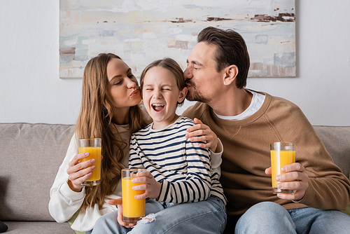 parents holding glasses of orange juice and kissing head of daughter at home