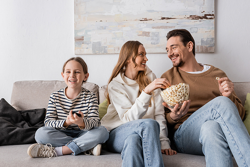 cheerful parents eating popcorn near happy daughter with remote controller