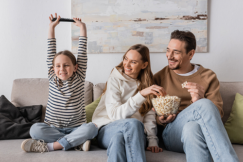 cheerful parents eating popcorn near excited daughter with remote controller