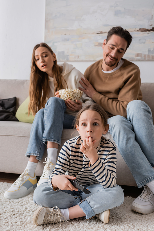 full length of scared kid watching movie near parents sitting with popcorn on blurred background