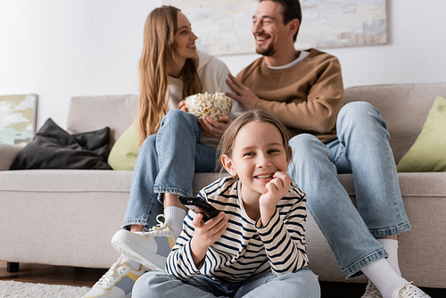 cheerful kid with remote controller watching movie near parents sitting with popcorn on blurred background