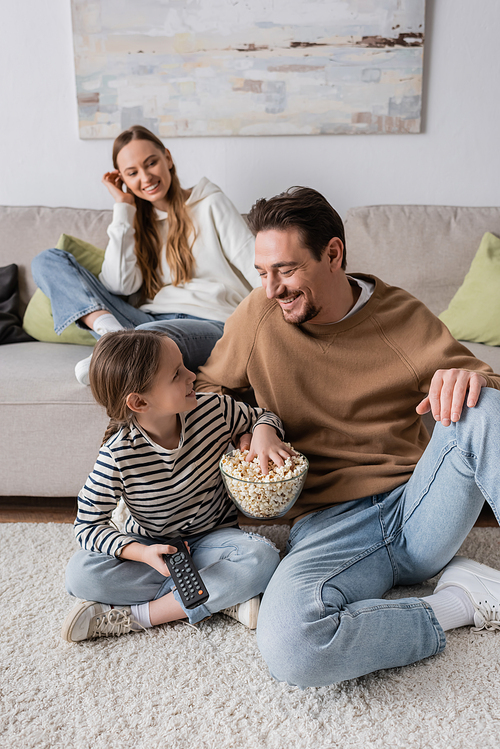 cheerful kid with remote controller looking at father while reaching popcorn near mother on blurred background