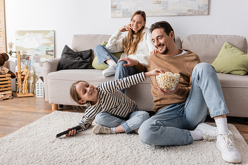 happy kid with remote controller reaching popcorn near father and mother on blurred background