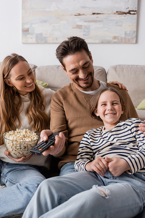 happy man holding remote controller while looking at daughter near wife with popcorn