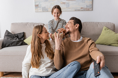 happy man holding remote controller and reaching popcorn near wife and cheerful daughter on sofa