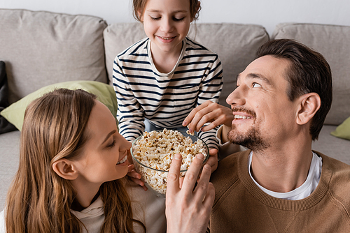 happy man reaching popcorn near wife and cheerful daughter on sofa