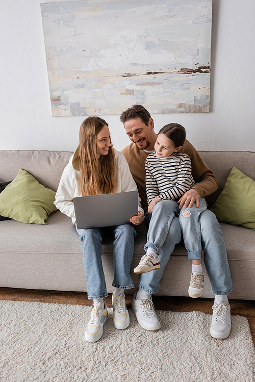full length of happy woman sitting with laptop near husband and daughter in living room