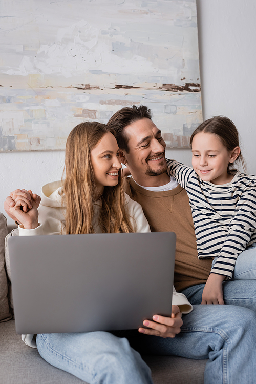 happy parents holding hands near daughter looking at laptop in living room
