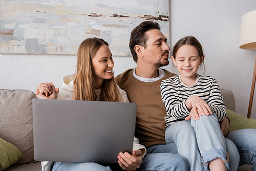 happy woman sitting with laptop near husband kissing daughter in living room