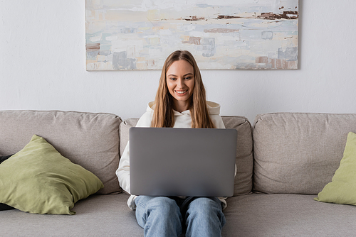 cheerful woman in casual clothes using laptop while sitting on couch