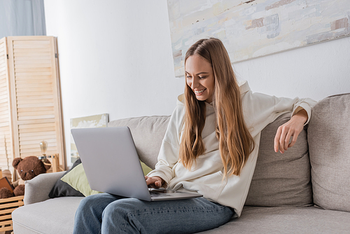 positive freelancer in casual clothes using laptop while sitting on couch