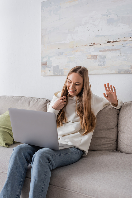 positive freelancer in casual clothes waving hand while using laptop in living room