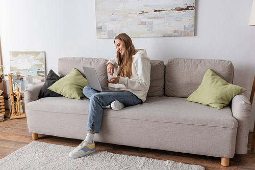 full length of positive woman in casual clothes having video call on laptop in living room