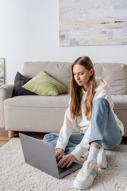 woman in casual clothes sitting on carpet near couch and using laptop