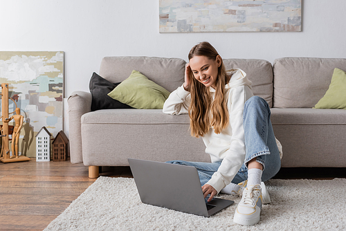 cheerful woman in casual clothes sitting on carpet near couch and using laptop