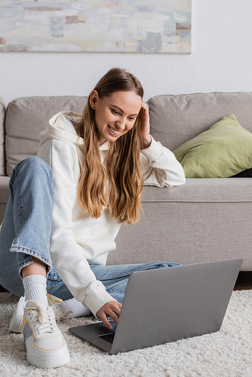 positive woman in casual clothes sitting on carpet near couch and using laptop