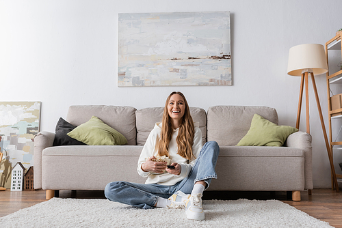 full length of joyful woman holding remote controller while watching movie in living room