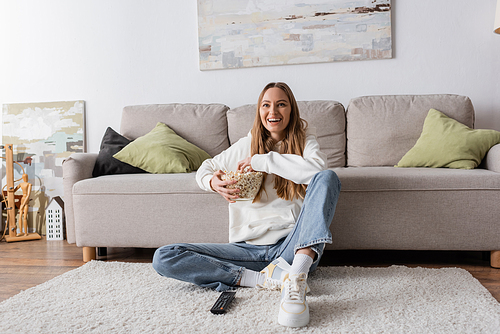 full length of joyful woman holding bowl with popcorn while watching movie in living room