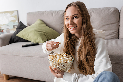 happy woman holding bowl with popcorn while watching movie in living room