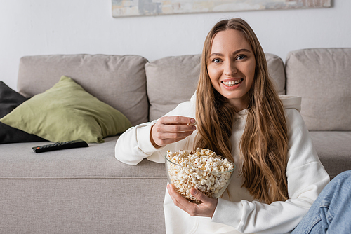 cheerful woman holding bowl with popcorn while watching movie in living room