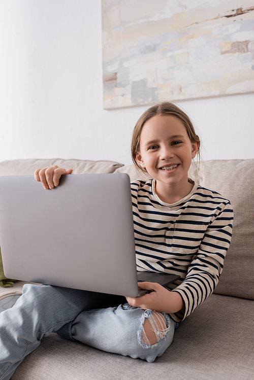 joyful preteen girl in casual clothes looking at camera and holding laptop while sitting on couch