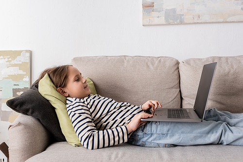 side view of cheerful preteen girl in casual clothes using laptop while lying on couch