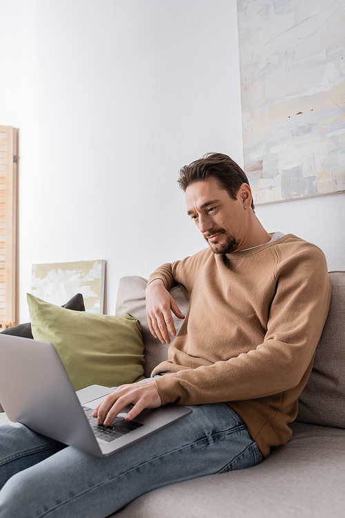 bearded man using laptop while sitting on sofa in living room
