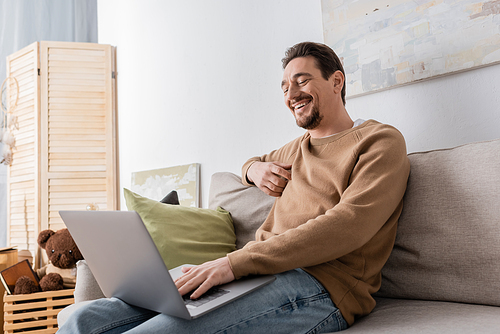 cheerful man using laptop while sitting on sofa in living room