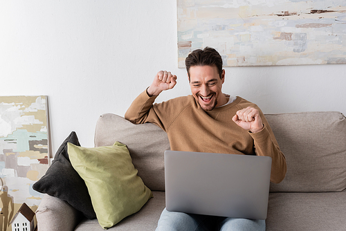 positive man watching championship on laptop while cheering and sitting on sofa in living room