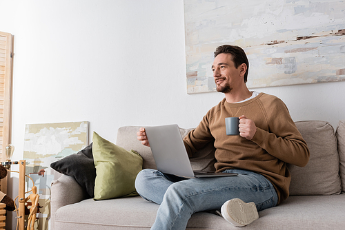 smiling man using laptop while holding cup and sitting on sofa in living room