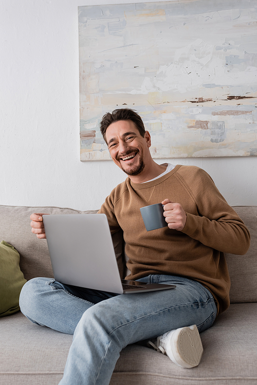 joyful man using laptop while holding cup and sitting on sofa in living room