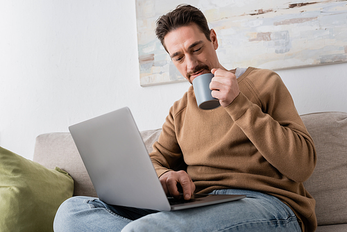 bearded man using laptop while holding cup of coffee and sitting on sofa in living room