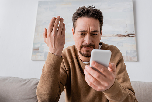 low angle view of displeased man in beige jumper looking at smartphone while gesturing at home