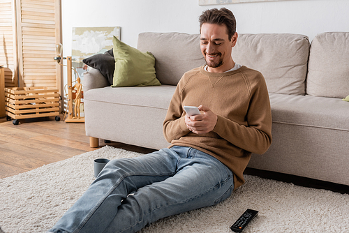 happy man in beige jumper messaging on smartphone while sitting on carpet at home