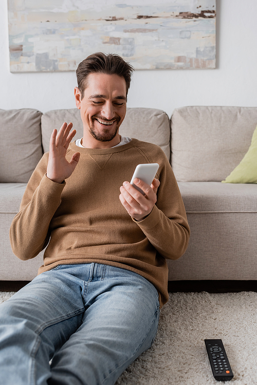 happy man in beige jumper waving hand at smartphone while having video call at home