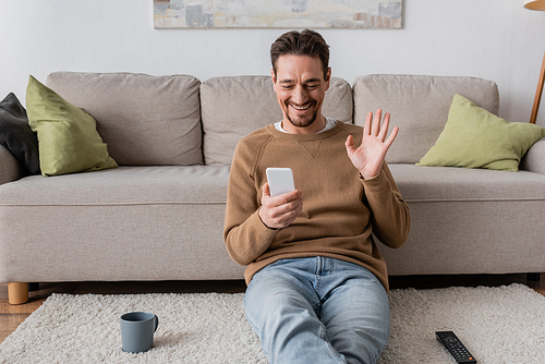 cheerful man in beige jumper waving hand at smartphone while having video call at home